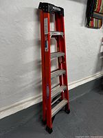 Photo of a 6 foot red aluminum folding ladder with black top hinges and rubber feet, leaning against a wall in a basement-like space.