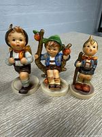 Three Hummel figurines showing children in different poses: one standing with two rabbits in a basket, one holding a pumpkin, one seated on a branch with apples