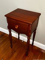 Antique maple night stand shown from a side angle against a grey wall, displaying the single drawer, turned spindle decorations, and tall turned legs.