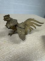 Photo showing a brass rooster figurine with detailed feathers and tail, placed on a wooden surface.