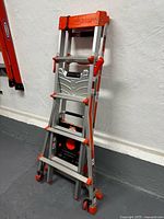 Photo of an aluminum Little Giant folding ladder in a compact folded position showing orange locks and wheels.