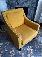 Photo showing a gold/yellow upholstered living room or bedroom chair with silver nailhead trim. The chair is placed on a patterned rug and is in front of a fireplace.