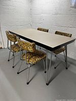 Full view of the white Formica top dining table with 4 floral upholstered chairs around it against a concrete wall.