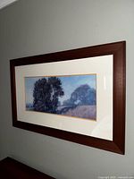 Framed landscape wall art depicting a country hill scene with large trees and misty background, enclosed in wood frame under glass.