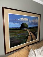 Framed wall art showing a country road landscape with a tree and clouds, hung on a wall above a couch.