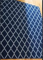 Photo of a blue area rug with a gray diamond lattice pattern placed on a wooden floor. The rug's texture and pattern are clearly visible.