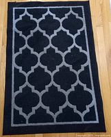 Top-down view of the black and grey rectangular area rug with geometric trellis design on wood floor.