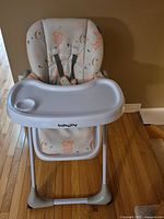 Front view of BabyJoy baby high chair showing padded seat with animal print cushion and attached white tray with cup holder.