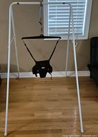Full view of white metal frame stand with black harness suspended from a spring for baby jumping.