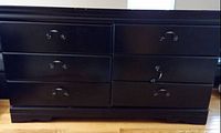 Front view of a sturdy black wooden dresser showing six drawers with metal handles. Dresser is placed on light wooden flooring.