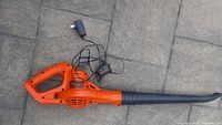 Top view of the orange and black Black & Decker cordless leaf blower with charger cable placed on a stone pavement.