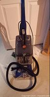 Front view of Hoover carpet cleaner showing upright form, hose coiled at base, and cleaning solution tank.
