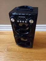 Front view of black Sylvania portable house speaker sitting on wooden floor showing dual speaker units, control knobs, buttons, and top handle.