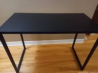Black rectangular office table with metal frame legs on a hardwood floor against a beige wall. Table surface flat and clean.