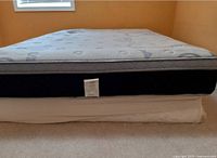 Side view of queen mattress on white bed slate, showing mattress label attached to black side panel with patterned light top surface. Room features tan walls and carpeted floor.