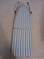 Top view of small portable ironing board showing blue and white striped fabric cover stretched over padded surface, white metal frame partially visible.