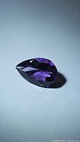 Pear-shaped purple amethyst gemstone showing facets and deep purple color under light on a plain surface.