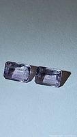 Pair of natural amethyst gemstones, emerald cut, light purple color, placed on white background.