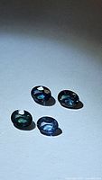 Four oval cut blue sapphires with green undertones placed on a flat surface under natural lighting, showing facets and clarity.