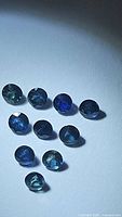 Ten loose round cut blue sapphires laid out on a white surface, showing their blue color and size approximately 3mm each.