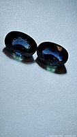 Pair of deep blue oval cut sapphires shown side by side on a flat surface, highlighting size and color under direct light.