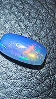 Close-up photo of a long cushion-shaped natural Ethiopian crystal opal cabochon showing vibrant blue and multicolored flashes on a dark textured surface.