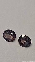 Two oval faceted sapphires in natural light showing dark purple and grayish bluish color tones.