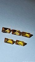 Five bright yellow-orange, baguette cut sapphires arranged on a flat surface showing clarity and color under direct lighting.