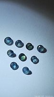 Ten small round multicolored blue and green natural sapphire gemstones arranged loosely on a white background under direct light showing their faceted surfaces and color variation.