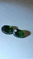 Two vivid green natural Tsavorite garnet gemstones, oval cut, displayed on a plain surface under light showing color and facets clearly.