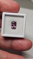 Cushion-cut pink spinel gemstone displayed in white foam within a small display box, held by fingers.
