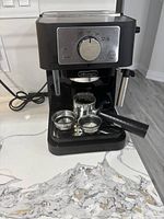 Photo showing the front of the black and silver DeLonghi espresso machine with manual control knob and accessories on the drip tray: portafilter, filter baskets, tamper.