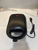 Closed black waffle maker with attached power cord and Type B plug on kitchen counter