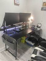 Gaming corner setup showing black metal framed desk with dual mounted monitors and side storage pouch, with various gaming accessories on the desk.