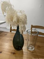 Tall dark green glass vase with pampas grass and smaller clear glass vase side by side on wooden table