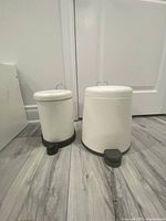 Two off-white plastic step trash cans with black foot pedals and metal handles shown from front and side angles on a gray wood floor against a white wall.