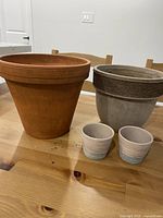 Four clay pots arranged on a wooden table: one large terracotta, one medium gray, and two small cream-colored ribbed pots.