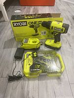 Ryobi ONE+ drill, battery, charger, 65 bit set in case, and box arranged on wooden floor.
