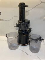 The Tuumist electric juicer in black with two clear plastic collection cups placed in front, showing the complete lot contents.