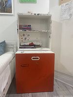 Front view of wooden rolling storage cabinet showing white open shelving and large red drawer with marks and writing.