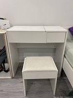 White wood composite vanity table with matching stool. The vanity shows a simple clean-lined design and white finish. Stool has padded seat.