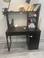 Front view of black wood desk with built-in shelving holding books and small items, drawer and cabinet visible on right side.