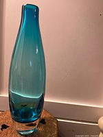 Tall vintage blue blown glass vase with clear thick base, showing gradient blue color, displayed on wooden surface against pale pink wall.