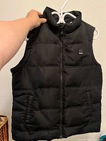 Front view of black sleeveless puffer vest with zipper and pockets, brand label on chest.