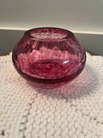 Small rounded cranberry coloured blown glass vase with textured internal pattern, shown from one angle on a white knitted surface.