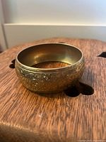 Close-up photo of the brass bangle showing intricate floral engravings on the curved surface, placed on a wooden surface.