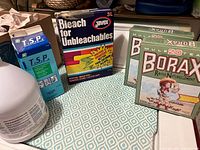 Photo shows various cleaning products including borax, Javex bleach, TSP powder box and bottle on patterned blue and white tablecloth.