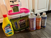 Photo showing all cleaning supplies together including Pink Solution large bucket with pink lid, small pink soap container, Javex multi gel bleach bottle, and three clear bottles of Castile liquid soap in lavender, orange, and blue colors.