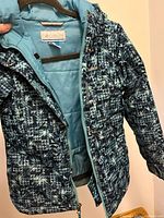 Front view of the blue geometric patterned Columbia winter jacket hung on a hanger, showing zipper and snap closures.