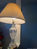 Photo of entire crystal table lamp lit, showing beige pleated fabric lampshade and clear crystal base with intricate cut pattern.
